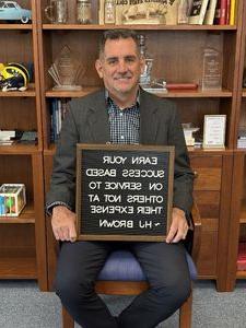 a photograph of Phil Douthitt sitting in a chair and holding a board which displays the quote "EARN YOUR SUCCESS BASED ON SERVICE TO OTHERS NOT AT THEIR EXPENSE"